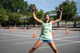 A student in a light green shirt and blue shorts jumps in the air with arms raised, holding a drink and a smartphone in an empty parking lot marked with orange cones.