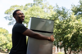 A smiling young man in a black t-shirt carries a stainless steel Whirlpool mini-fridge outdoors with green trees and a college building in the background.