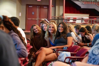 Four female students seated in a red auditorium, smiling at the camera