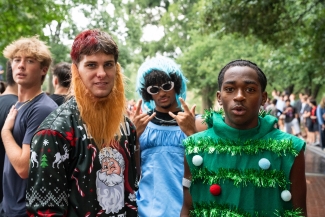 Three young men pose together in festive costumes, including a holiday sweater with a false beard, a blue dress with a wig, and a green Christmas tree tunic.