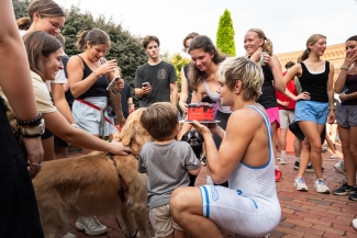 A crouched runner in a white singlet holds up a small red cake to show a young child surrounded by a crowd of people and dogs on a brick patio.