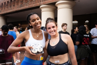 Two smiling young women in athletic sports bras pose together outside, with one holding a plastic container of four iced cupcakes.