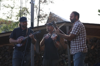 A three-piece acoustic band performs outdoors, playing a mandolin, a violin, and an acoustic guitar in front of stacked firewood.
