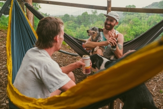 Two men relax outdoors in hammocks, holding canned drinks while one holds a blue heeler dog on his lap.