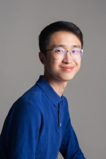 A male student with glasses and a blue button down smiles softly at the camera with head turned