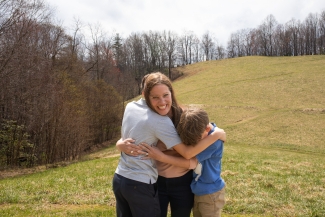 A smiling woman standing on a grassy hill outdoor field hugs two young boys tight against her.