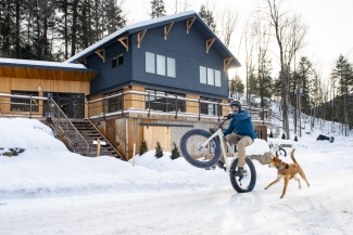 A cyclist pops a wheelie on a fat-tire bike on a snowy road outside a two-story wooden lodge while a dog runs alongside.