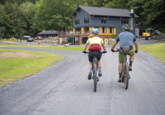Two cyclists ride mountain bikes side by side down a gravel driveway toward a large blue lodge.