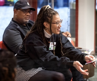 A woman with dreadlocks and glasses gestures forward with open hands while seated next to a man during an indoor discussion.