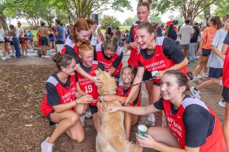 A group of female athletes in red Davidson Lacrosse pinnies kneel together on a brick walkway to pet a golden retriever.