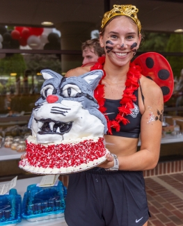 A smiling participant in a tiara and ladybug wings holds a large cake decorated to look like a grey and white wildcat head.