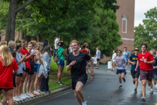 A smiling male runner in all-black athletic wear leads a pack of participants down a wet campus road while spectators clap from the curb.
