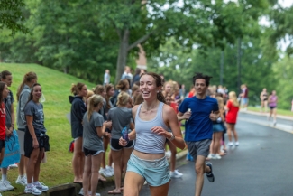 A smiling female runner in a white tank top and light blue shorts leads a group of participants down a paved road lined with spectators.