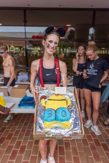A student wearing mouse ears and red suspenders holds a cardboard box containing a custom yellow cake decorated to look like a Minion character.