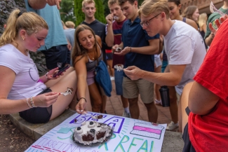 A group of young adults gather around a decorated sign on the ground, using plastic forks to sample a cookies-and-cream cake.