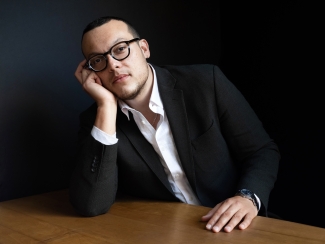 Portrait of poet Charif Shanahan wearing glasses and a dark suit jacket, leaning his head on his hand at a wooden table against a dark background.
