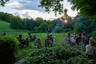 A group of people sits in lawn chairs outdoors watching a speaker near a campfire during sunset.