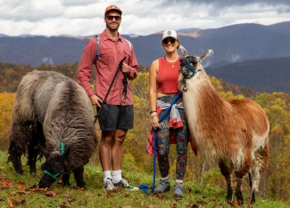 A man and woman wearing hats stand side-by-side with two leashed llamas on a grassy hilltop against mountain views.