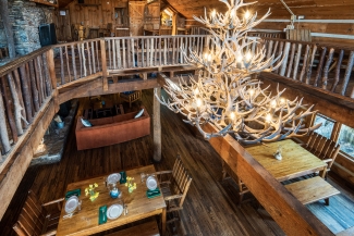 High-angle view of a rustic wooden dining hall featuring set dining tables and a large antler chandelier hanging from the vaulted ceiling.