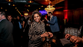 A smiling woman wearing a zebra-print top holds a drink while standing at a dark venue bar.