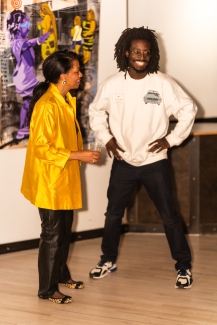 A woman in a bright yellow jacket talks with a man in a white sweatshirt at an art gallery event.