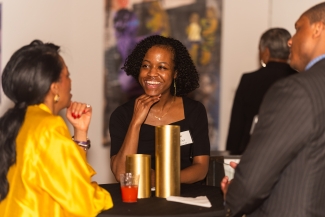 A woman in a black top smiles while conversing with others at a high-top table during an evening event.