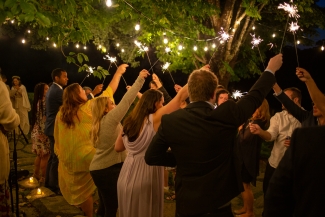 Wedding guests outdoors at night celebrate under string lights while raising glowing sparklers into the air.
