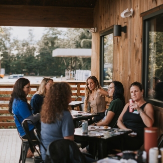 A group of women sits around small black tables chatting on a covered wooden porch outdoor patio.