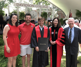 A group of five people standing together outdoors in front of a "Davidson College" sign. Center stage is a smiling graduate in a black gown and red stole holding a diploma folder, flanked by supporters and President Emerita Carol Quillen in red academic regalia.