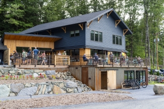 Guests relax and dine on the elevated wooden deck of a modern blue and wood-paneled lodge.