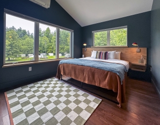 A modern bedroom features a large bed with colorful linens, dark walls, a green-and-white checkered rug, and large windows overlooking trees.