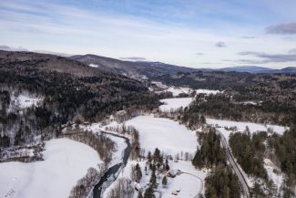 An aerial view of a snow-covered mountain valley with a winding river, dense evergreen forests, and roads running through snow-covered fields.