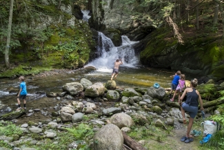 A person leaps into a natural swimming hole at the base of a multi-tiered waterfall surrounded by mossy rocks and forest greenery.