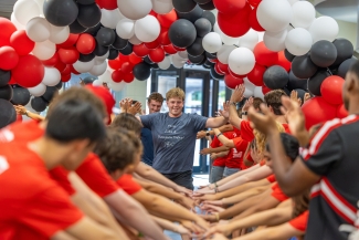 A young man walking down a hallway filled with red, white, and black balloons while crowd members in matching red shirts cheer and high-five him.