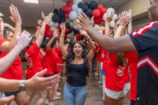 A young woman in a black tank top and jeans runs down a hallway filled with red, white, and black balloons while crowd members in matching red shirts cheer and high-five him.
