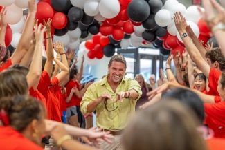 A smiling parent in a green shirt runs through the balloon tunnel full of smiling, cheering students
