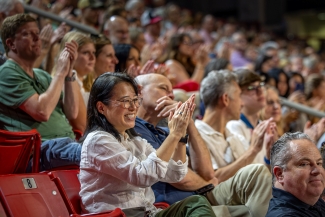 A smiling parent in a blouse and glasses claps, seated in the stands in Baker Sports arena
