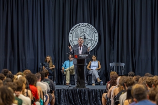 President Doug Hicks onstage in front of a Davidson College seal and black backdrop, addressing a crowd of new students