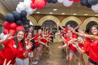 A cheering group of orientation leaders in matching red shirts reaching out to high-five incoming students beneath a balloon archway.