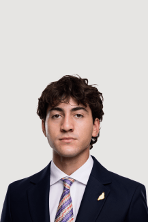 A male student in a suit and pink striped tie with dark curly hair and a neutral expression