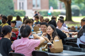 Students and families sit together at outdoor picnic tables, eating lunch from boxed meals during a campus orientation event.