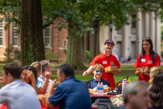 Orientation team members in red shirts hand out boxed lunches to students and families seated outdoors at picnic tables.