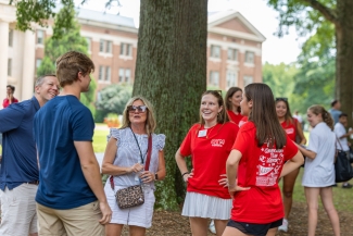 A group of students and parents stand near a large tree on campus, conversing with orientation team members dressed in matching red shirts.