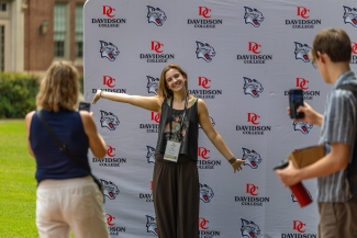 A student poses with arms outstretched in front of a Davidson College backdrop while people take photos of her with their smartphones.
