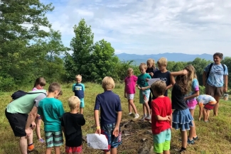 A group of children and adults gather outdoors in a grassy field holding maps for an activity.