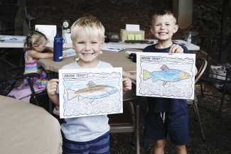 Two smiling young boys hold up completed coloring pages depicting colorful brook trout.