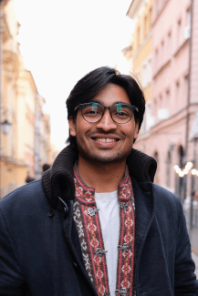 A male student wtih a navy blue jacket and colorful shirt, glasses, standing on a city street