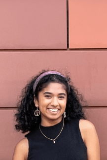 A woman with dark curly, shoulder-length hair in a purple headband stands in front of a red terra cotta wall