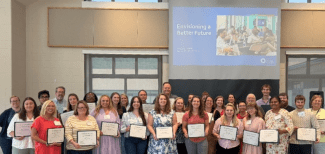 A group of award recipients stands together holding framed certificates in front of a presentation slide titled "Envisioning a Better Future."