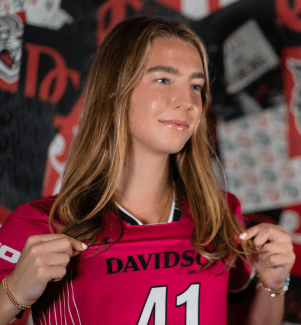 A young woman with long blonde hair smiles while holding out the collar of her pink Davidson soccer jersey featuring number 41